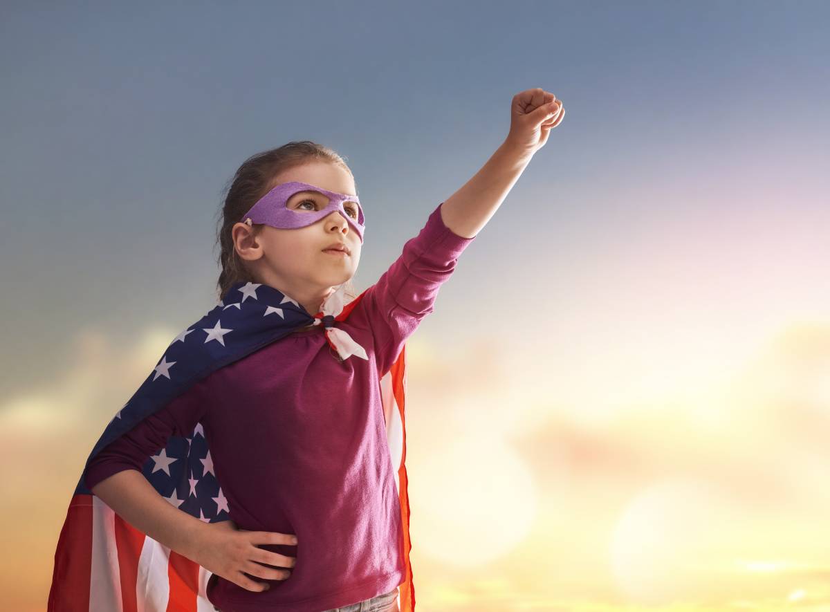 Child dressed up as superhero, wearing an American Flag cape with her hand in the air