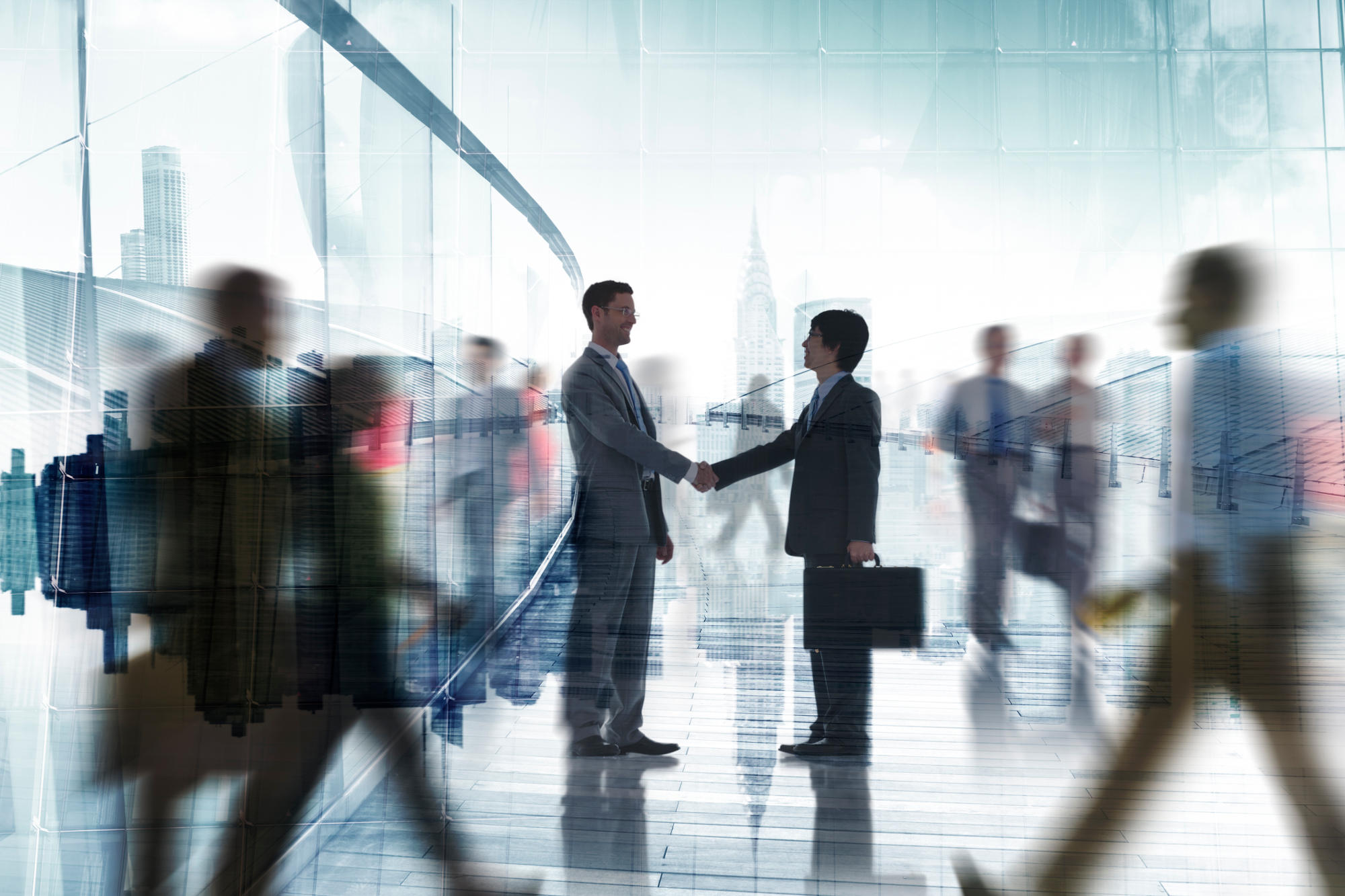 Two business people shaking hands with blurred people walking around them
