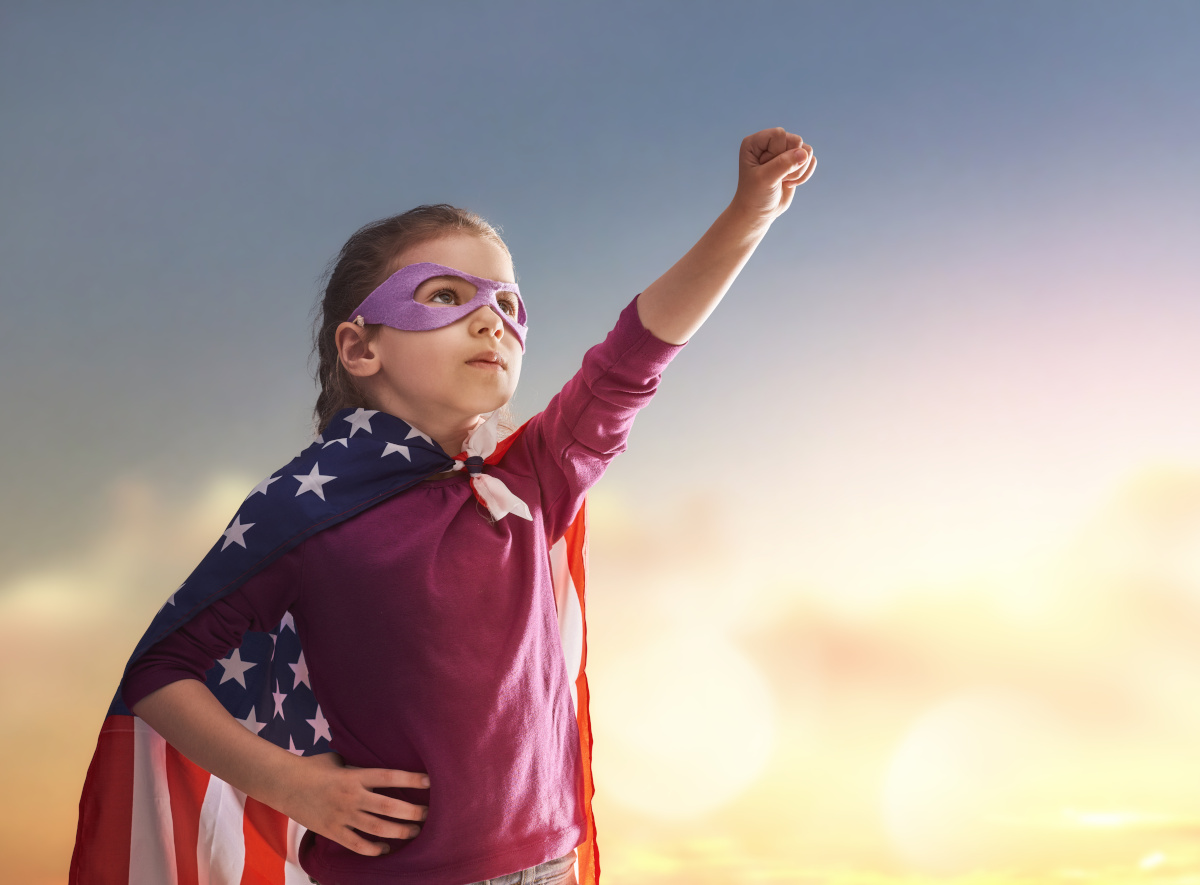 A young child wearing a mask and an American flag as a cape striking a superhero pose.