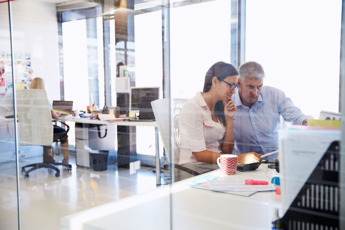 Male and Female Colleagues Working Together at Desk with Woman at Another Desk in Background